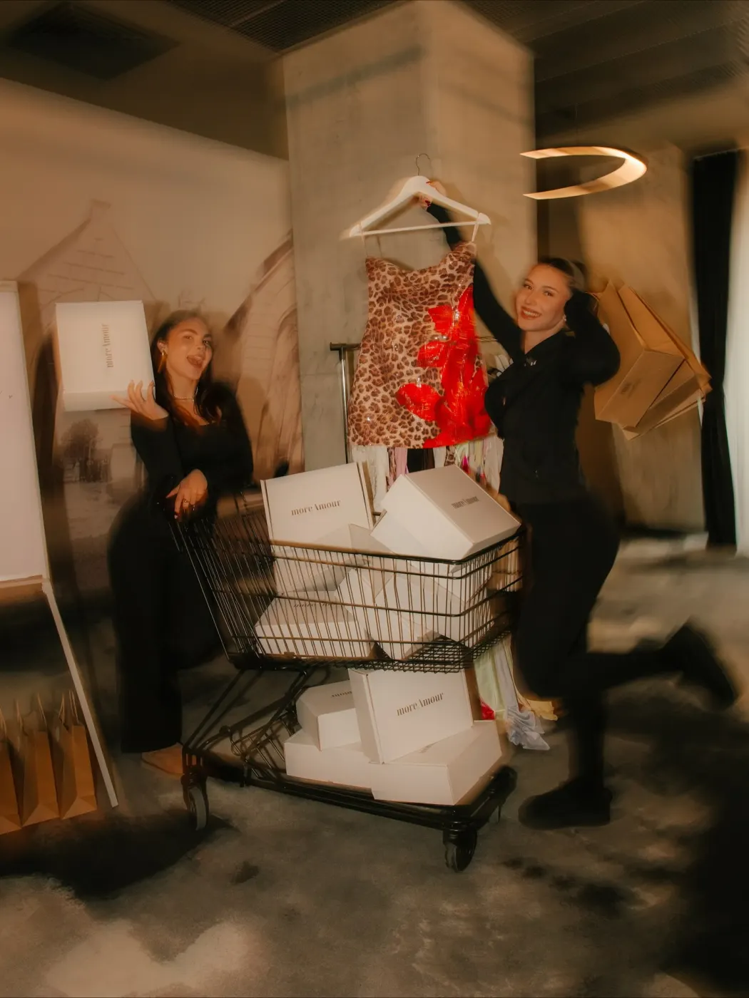 Două prietene lângă căruciorul plin de cutii moreAmour, una ridicând o rochie cu leopard