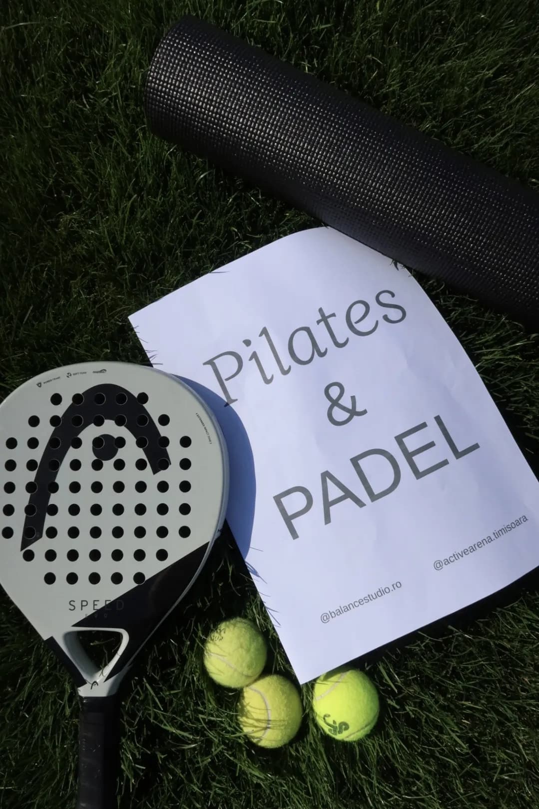 Rachetă de padel, saltea și mingi pe iarbă, lângă afișul Pilates & Padel